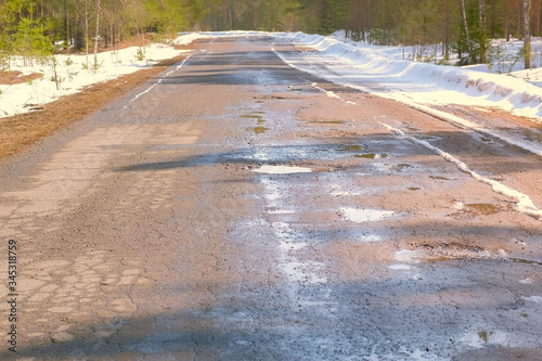 Destroyed broken asphalt country road with holes, melting snow and puddles early spring. Old village road after winter. Cracked ruined bad rural asphalt in forest, national park.