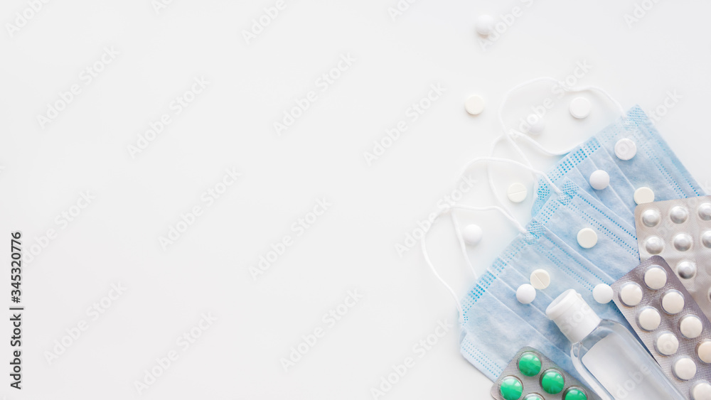 Top view on pack of blue protective medical masks, transparent bottle of sanitizer gel and different scattering pills. Coronavirus COVID-19 concept on  white background with copy space.