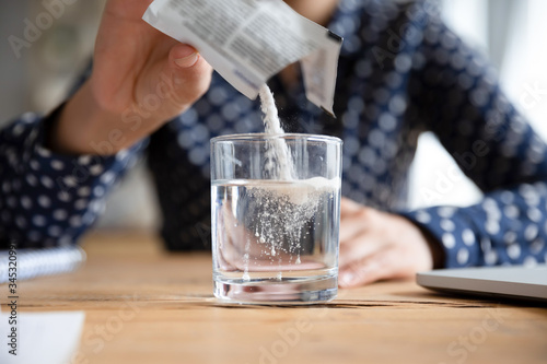Close up young indian woman pouring soluble anti-influenza powder in glass of water. Sick millennial girl student feeling unwell, relieving grippe fever flu symptoms, high temperature, headache. photo