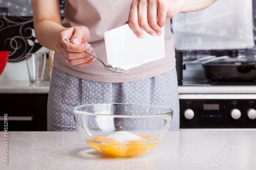 Female hands hold bag of vanilla in their hands and pour it into a spoon to add to the kneading dough - beaten eggs with flour and sugar against the background of kitchen utensils. Cooking pie at home