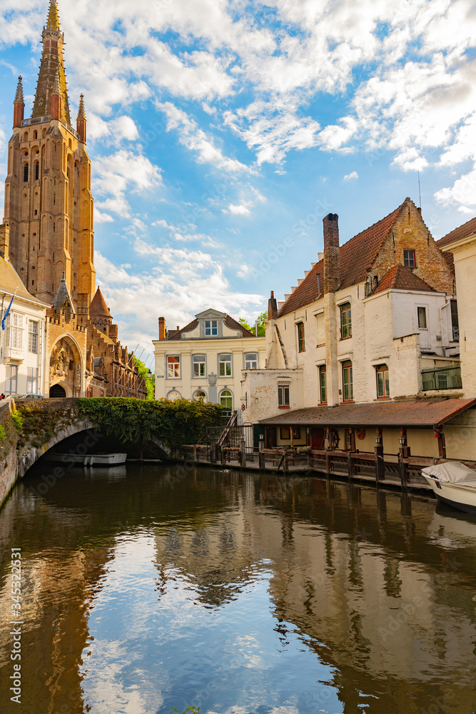 View of Dijver canal with canal boat, vine covered bridge, traditional buildings and Notre Dame Cathedral in Bruges, Belgium