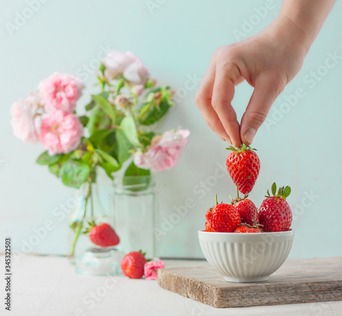 Woman takes or puts one strowberry in fruit bowl full of fresh, red, jucy strawberries. Pastel-colored decoration with a floral  arrangement against mint color background photo