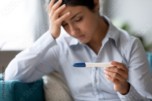 Focus on pregnancy test in hands of frustrated stressed young indian woman. Unhappy depressed millennial hindu girl dissatisfied with result, unwanted pregnancy, fertility problem, bad news concept. photo
