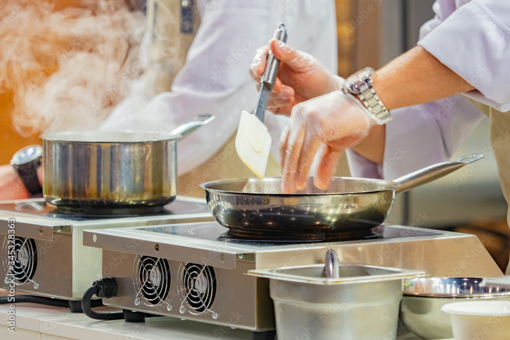Two chefs work in the kitchen of the restaurant. Cook fried foods in a pan. Heat treatment. A ladle and a frying pan in stainless steel. Cook's job. Equipment for cafes and restaurants.