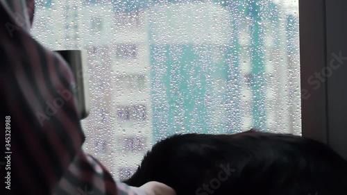 woman in sleepwear drinking coffee standing looking at rainy blue droplets on window stroking cuddling black cat pet laying on sill. quarantine self isolation at home. rainy stormy weather.