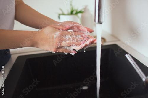 Young woman in kitchen during quarantine. Cut view of caucasian's hands in white soap bubbles hold sponge. Washing hands.