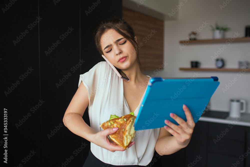 Young woman in kitchen during quarantine. Businesswoman talking on phone. Hold big tablet and sandwich in hands. Home office and remote work. Lunchtime period.