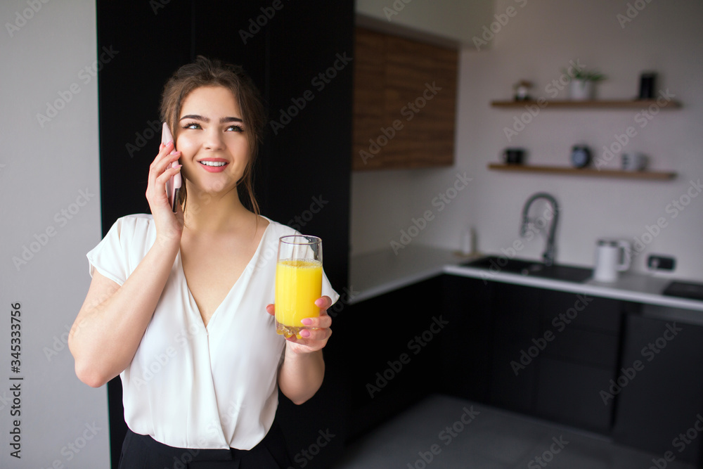 Young woman in kitchen during quarantine. Girl talking on phone and smile. Businesswoman hold glass of orange juice. Online wireless conversation. Young woman in kitchen during quarantine. Girl talking on phone and smile. Businesswoman hold glass of orange juice. Online wireless conversation.