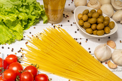 On a white wooden table, spaghetti, tomatoes, herbs, peppers, olive oil, garlic, mushrooms and olives are spread out in a white glass bowl.