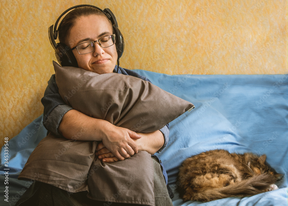 Fototapeta premium girl hugs a pillow and enjoy listening to favorite music with headphones while sitting on a blue sheet next to a sleeping cat