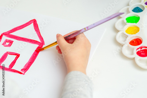 Child is drawing red house with watercolors on the white sheet of paper. Children creativity.