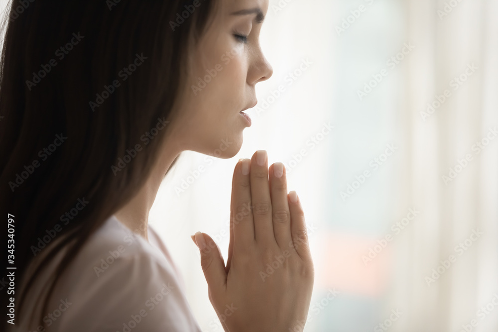 Close up side view of religious young woman hold hands in prayer asking God for good luck or fate, superstitious female believer pray at home, believe in future, faith, religion, superstition concept Close up side view of religious young woman hold hands in prayer asking God for good luck or fate, superstitious female believer pray at home, believe in future, faith, religion, superstition concept