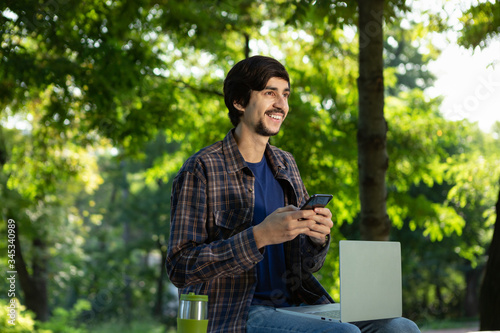 Young freelancer smiling sitting with a laptop in a park and holding smartphone. © ianachyrva
