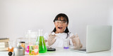 Photo of young adorable school girl holding a chemistry glassware while doing a scientific experiment and sitting at the modern white table with white laboratory wall as background.
