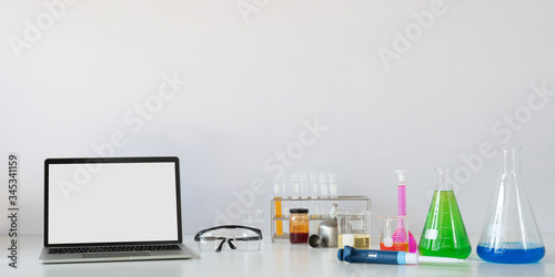 Slika na platnu Computer laptop with white blank screen putting on white working desk that surrounded by chemistry glass ware over laboratory white wall as background