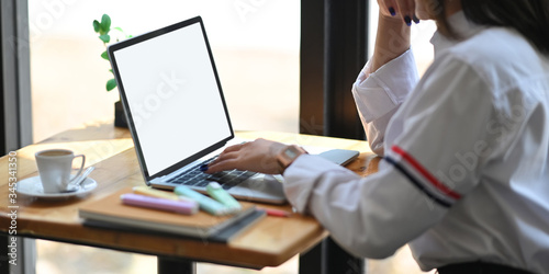 Cropped image of beautiful woman in white shirt keep hand on chin and  typing on computer laptop with white blank screen while sitting at the wooden working desk over office glass wall as background. © Prathankarnpap