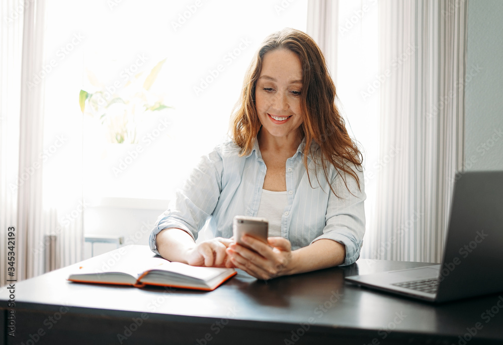 Adult smiling brunette woman doing notes in daily book and using the mobile phone with opened laptop at home Adult smiling brunette woman doing notes in daily book and using the mobile phone with opened laptop at home