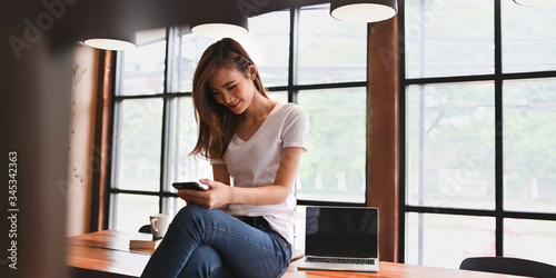 Foto Photo of beautiful stylish woman using a smartphone while sitting with crossed leg on the wooden long table over comfortable living room windows as background