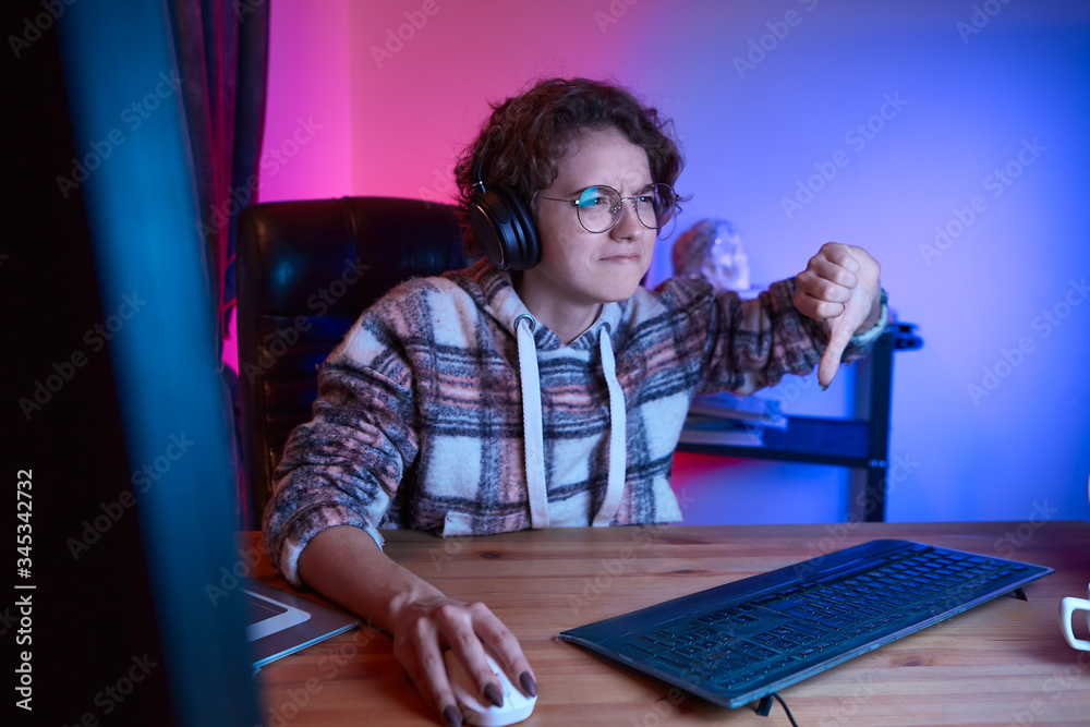 Upset girl holding thumb down looking at the screen of a gaming computer with weon light in the room Upset girl holding thumb down looking at the screen of a gaming computer with weon light in the room