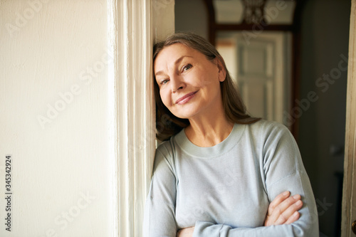 Attractive middle aged Caucasian woman with loose gray hair and wrinkles spending day indoors, leaning on white wall and smiling at camera. Mature people, aging, leisure and lifestyle concept