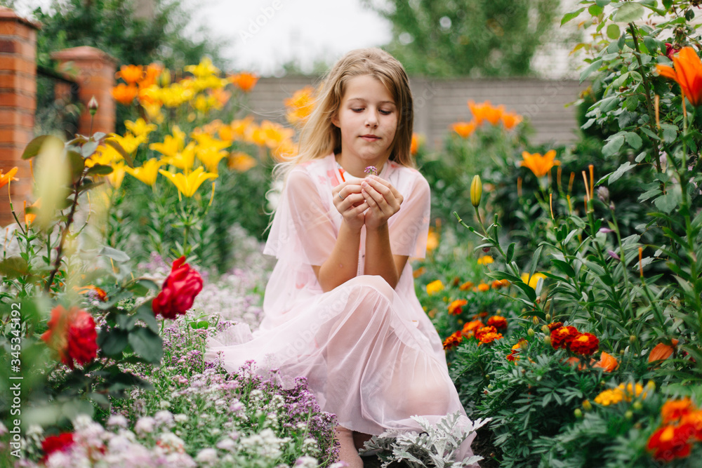 Naklejka premium beautiful blue-eyed girl with long blond hair. little girl in a pink flamingo dress. girl in the flower garden. summer bright, emotional photo. large, thick, bright flower garden near the house. 