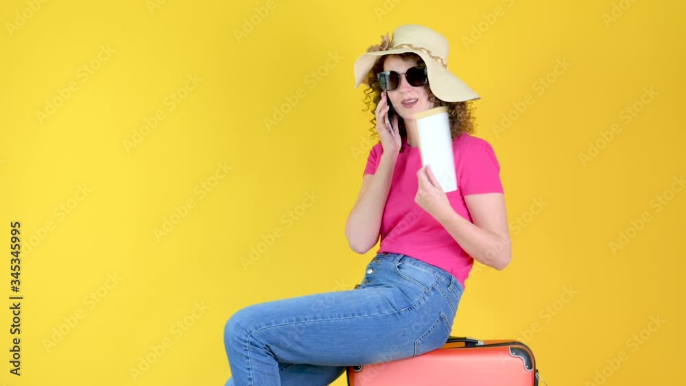 Attractive young girl in sunglasses sits on a suitcase with a ticket and uses a smartphone isolated over yellow orange background in studio. journey concept. Attractive young girl in sunglasses sits on a suitcase with a ticket and uses a smartphone isolated over yellow orange background in studio. journey concept.