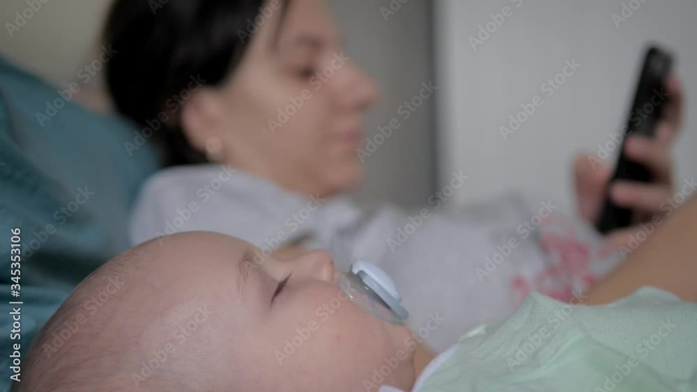 Baby Is Sleeping In Bed And Mother Is Near To Him Using A Smartphone Apps Baby Is Sleeping In Bed And Mother Is Near To Him Using A Smartphone Apps