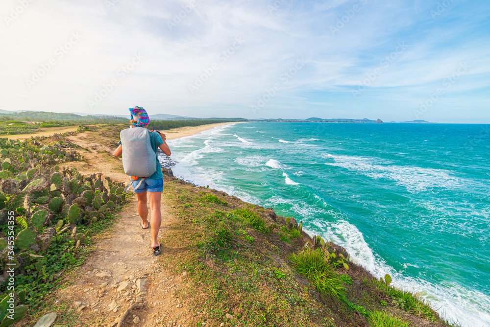 Woman with backpack looking at tropical coast from cliff above. Vietnam travel destination, Phu Yen province between Da Nang and Nha Trang. Gorgeous golden sand beach blue waving sea rock boulders,