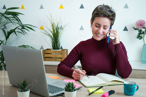 Woman talking on the phone while taking notes in her notebook next to a laptop. She is teleworking
 photo