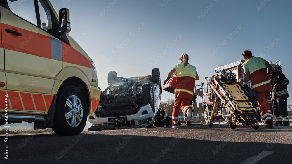 On the Car Crash Traffic Accident Scene: Team of Paramedics and Firefighters Rescue Injured People Trapped in Rollover Vehicle. Professionals Extricate Victims, give First Aid, Extinguish Fire