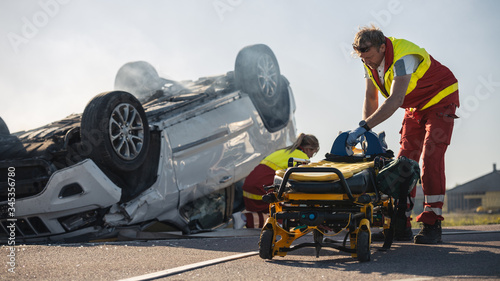 Fototapeta Naklejka Na Ścianę i Meble -  Paramedics and Firefighters Arrive On the Car Crash Traffic Accident Scene. Professionals Prepairing Stretchers For Rescue Injured Victim Trapped in Rollover Vehicle.
