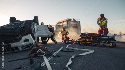 Paramedics and Firefighters Arrive On the Car Crash Traffic Accident Scene. Professionals Rescue Injured Victim Trapped in Rollover Vehicle by Extricating Them, giving First Aid and Extinguishing Fire photo