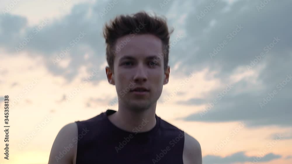Young man athlete in tank top running and looking at camera during sunset, close up slow motion Young man athlete in tank top running and looking at camera during sunset, close up slow motion
