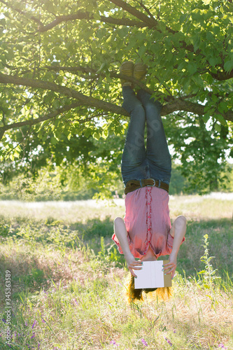 Quadro su tela Cheerful girl hanging on tree upside down and reading book