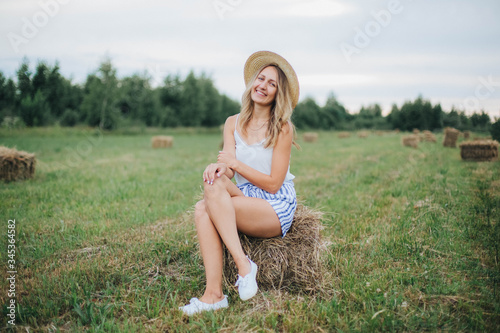 Fototapeta Naklejka Na Ścianę i Meble -  beautiful girl in a straw hat on a field with straw bales. girl sitting on the hay. blonde in the summer in a field with a hat. life in the countryside, outdoors. eco project. clean environment