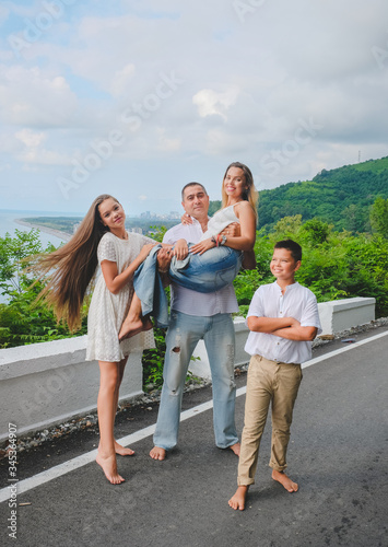 Cheerful, happy family, father, mother, son and daughter enjoying walk play outdoor near sea. Family in white clothes on walkway. Green forest background.