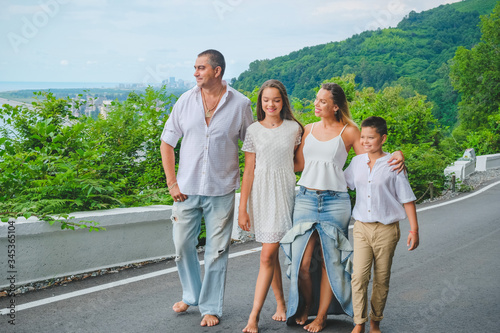 Cheerful, happy family, father, mother, son and daughter enjoying walk play outdoor near sea. Family in white clothes barefoot on walkway. Green forest background.