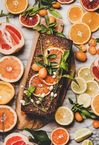 Citrus Mediterranean loaf cake. Flat-lay of almond pound cake with fresh oranges, lemon, kumquat, grapefruit and blossom flowers and leaves over grey marble background, top view. Vegan dessert concept