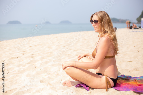 adult European woman with blond long hair sits on the sand by the sea in lotus position