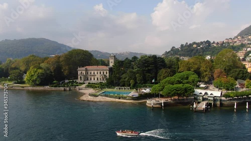 Aerial view of Villa Erba, pool, pier, motor boat sailing on Lake Como on a summer day amid mountains in a haze, green forest and Italian houses