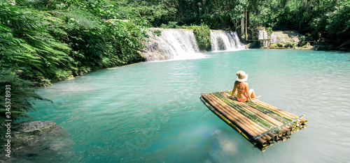 Woman in bikini and hat sitting on bamboo raft and enjoying view on waterfall. Travel and vacation concept. Banner and panoramic edition.