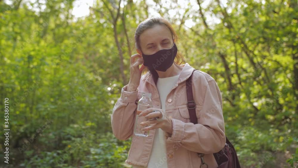 The woman removes the protective medical mask from her face to drink water. A woman walks alone in a Park during a coronavirus pandemic. The woman removes the protective medical mask from her face to drink water. A woman walks alone in a Park during a coronavirus pandemic.
