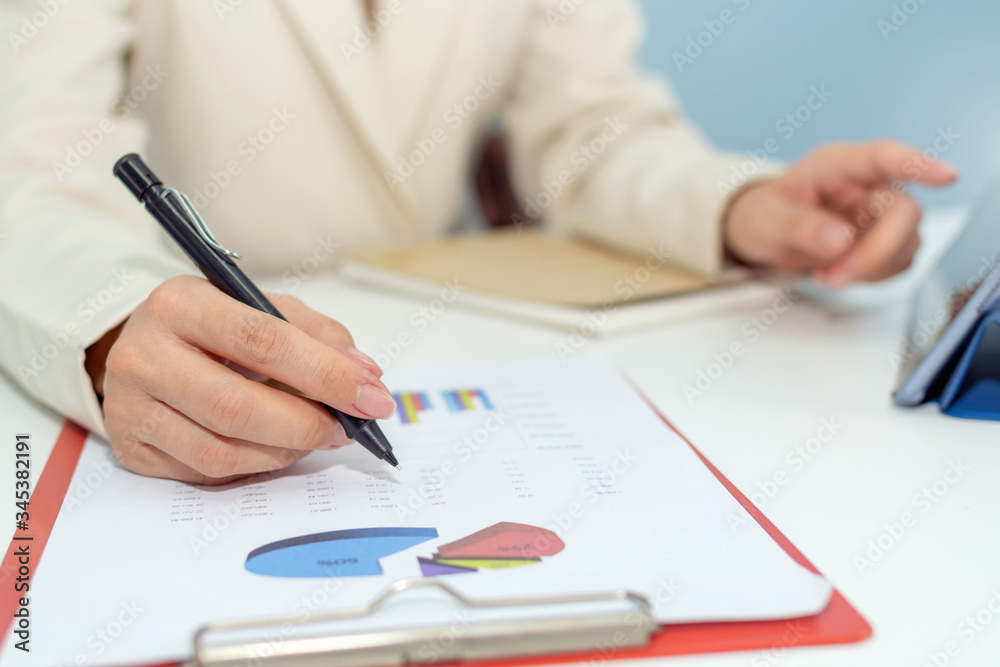 attractive businesswoman in white suit working and calculating about finance with strategy graph chart report and tablet on desk at office, digital online marketing and financial business concept