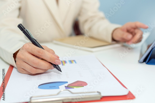 attractive businesswoman in white suit working and calculating about finance with strategy graph chart report and tablet on desk at office, digital online marketing and financial business concept