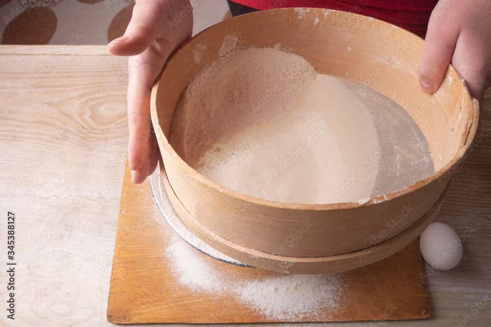Large bowl, cutting board, sieve for sifting flour on the table. Female hands sifting flour in the home kitchen. Large bowl, cutting board, sieve for sifting flour on the table. Female hands sifting flour in the home kitchen.