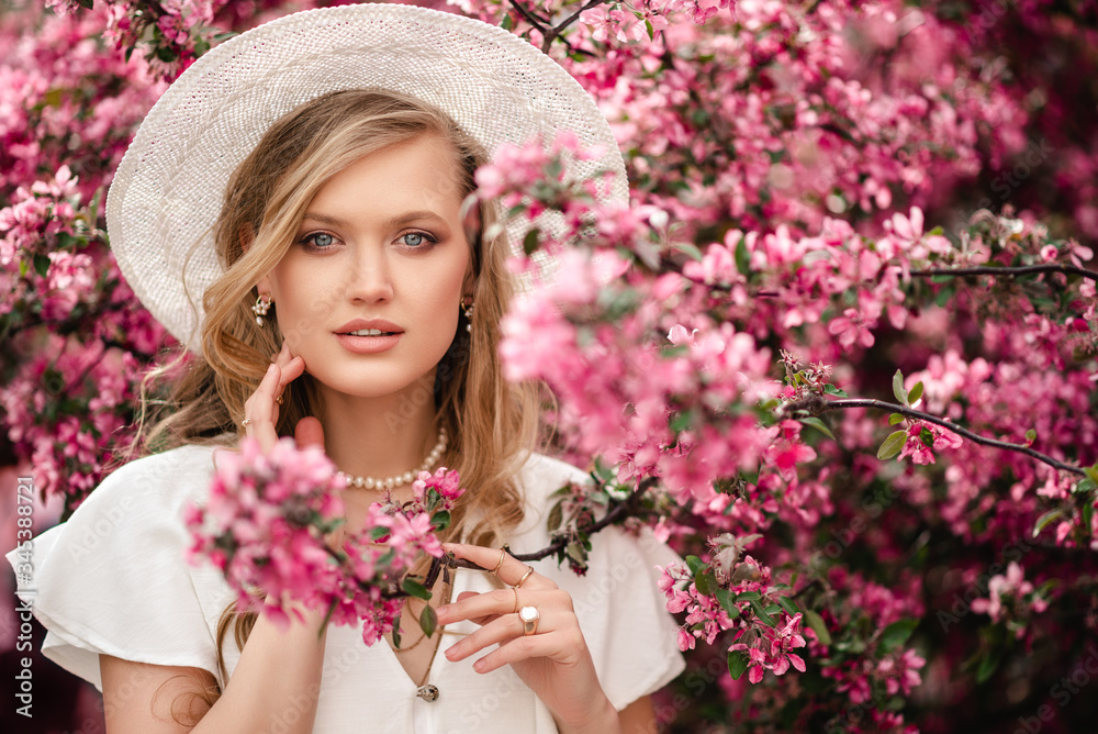 Young beautiful elegant lady wearing white wicker hat, pearl jewelries, dress, posing in pink spring blossom trees. Close up portrait. Copy, empty space for text