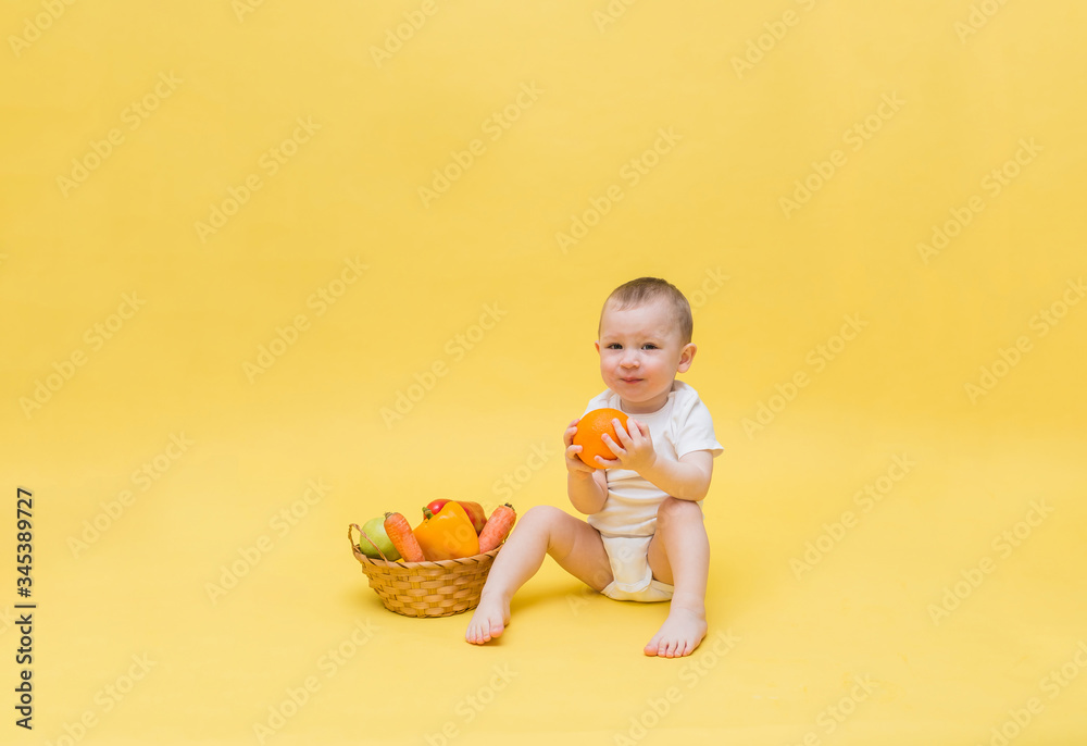 A cute boy is sitting in a white bodysuit and holding an orange. A small boy is sitting with a basket of vegetables and fruit on a yellow background. The kid looks at the camera. A cute boy is sitting in a white bodysuit and holding an orange. A small boy is sitting with a basket of vegetables and fruit on a yellow background. The kid looks at the camera.