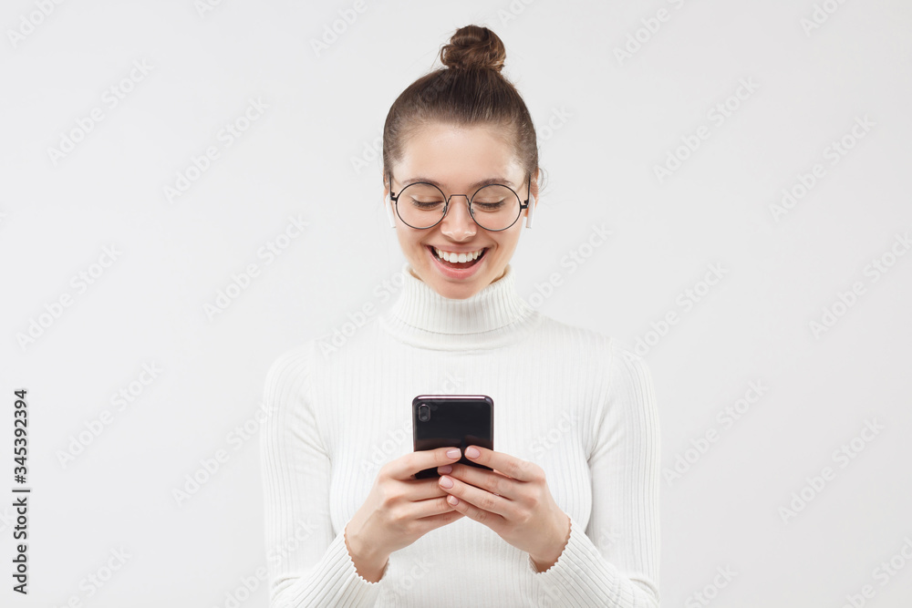 Young female in round glasses feeling relaxed and happy, laughing at funny content she is watching on smart phone screen, isoalated on gray background Young female in round glasses feeling relaxed and happy, laughing at funny content she is watching on smart phone screen, isoalated on gray background