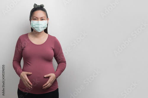 Asian pregnant woman wearing surgical mask looking at camera and holding her stomach by two hands, studio lighting portrait and copy space on grey background, covid19 concept