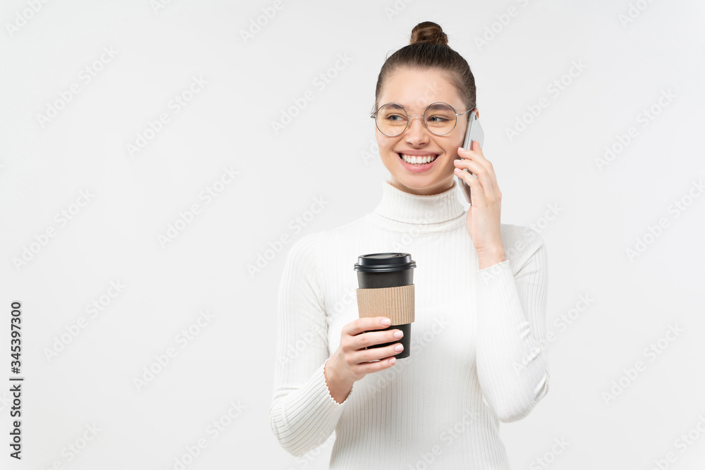 Young smiling woman talking on phone, laughing at jokes, holding coffee to go cup, isolated on gray background in studio with copy space Young smiling woman talking on phone, laughing at jokes, holding coffee to go cup, isolated on gray background in studio with copy space